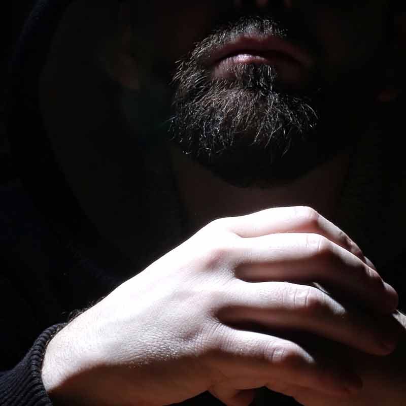 A bearded man shrouded in a hood holds his hands together.