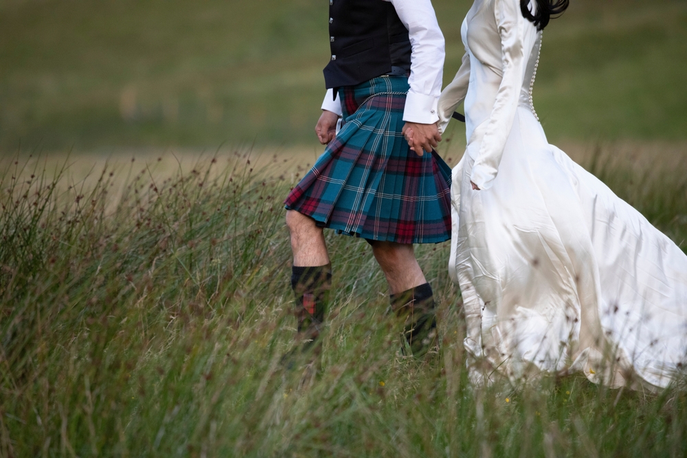 A newly married Scottish couple strolls through a grassy field in Glencoe, the Scottish highlands. He dressed in a Scottish Kilt and she dressed in a white wedding dress.