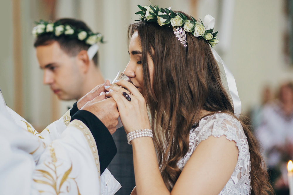 Stylish bride and groom in floral crowns drinking holy eucharist during matrimony in church. Wedding ceremony in cathedral. Classic spiritual wedding couple.