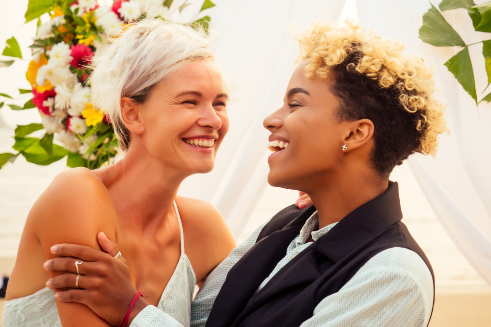 african american female groom in black suit and caucasian bride in dress in ceremony on tropical beach under wedding flover arch.