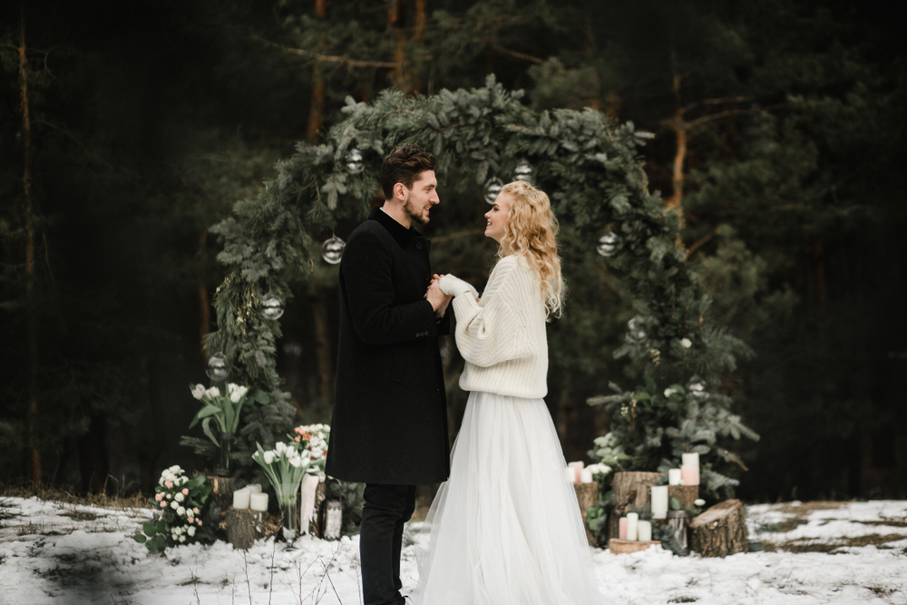 beautiful young wedding couple stands near the arch in the winter in the snow under the open sky at the ceremony and look at each other.
