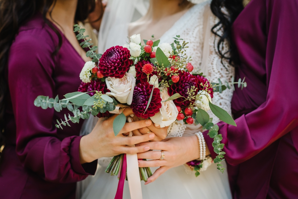 Bride and bridesmaids in elegant dress holding in hands bouquets of pastel burgundy flowers dahlia and greens eucalyptus with ribbon at nature. Young beautiful girls hold wedding bouquet outdoors.