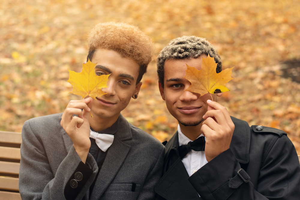 Young handsome black gay couple in love sitting on bench in park during autumn holding yellow leaves